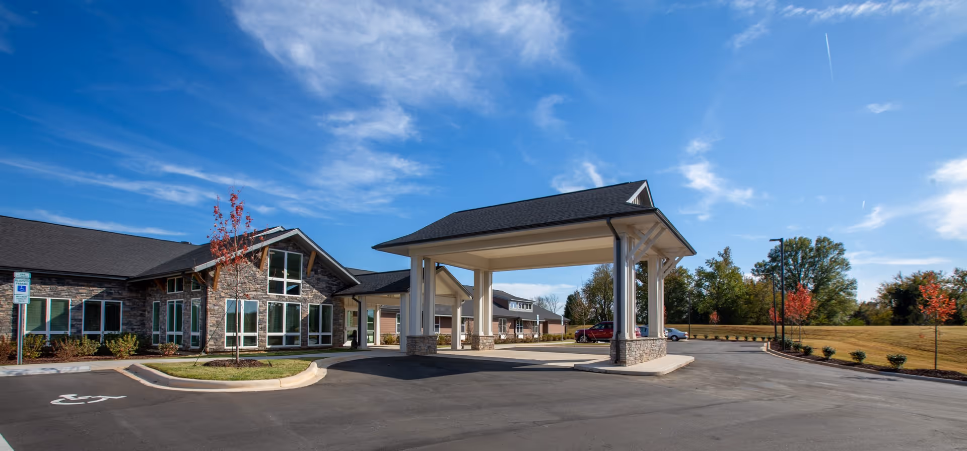 Exterior view of a senior living facility named The Landings of Cabarrus, showing a covered entrance with stone and white pillars, a parking area with a handicap parking space, and a building with large windows under a blue sky with some clouds.