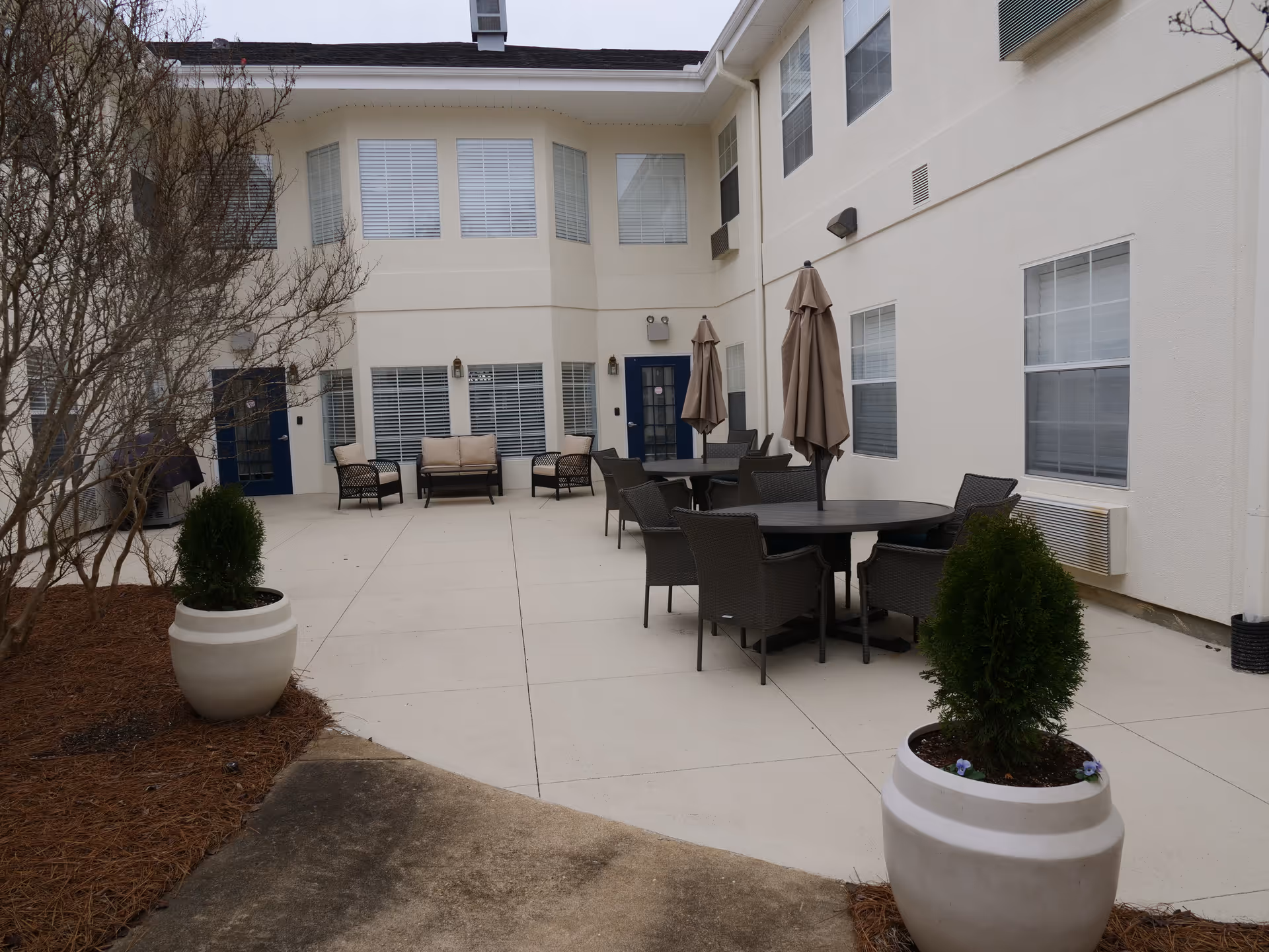 Outdoor courtyard area of a senior living facility with several round tables and chairs, each table having a closed umbrella. There are also cushioned chairs and a loveseat against the building wall. Two large white planters with small green shrubs are placed on the concrete ground near the seating area. The building has multiple windows with white blinds and beige walls.