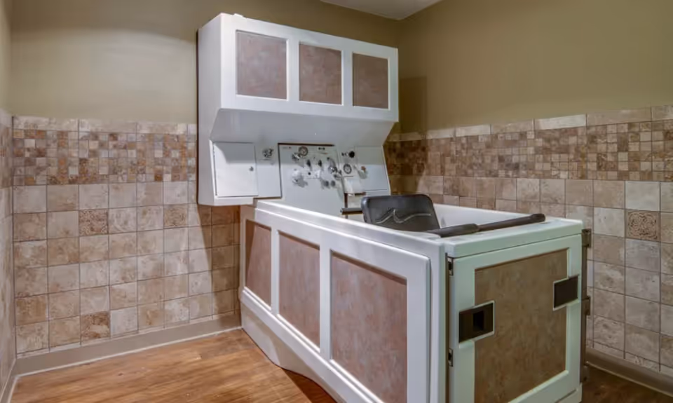 A specialized bathing tub designed for assisted bathing in a nursing center, featuring control knobs and a built-in seat, surrounded by beige and brown tiled walls and wood flooring.