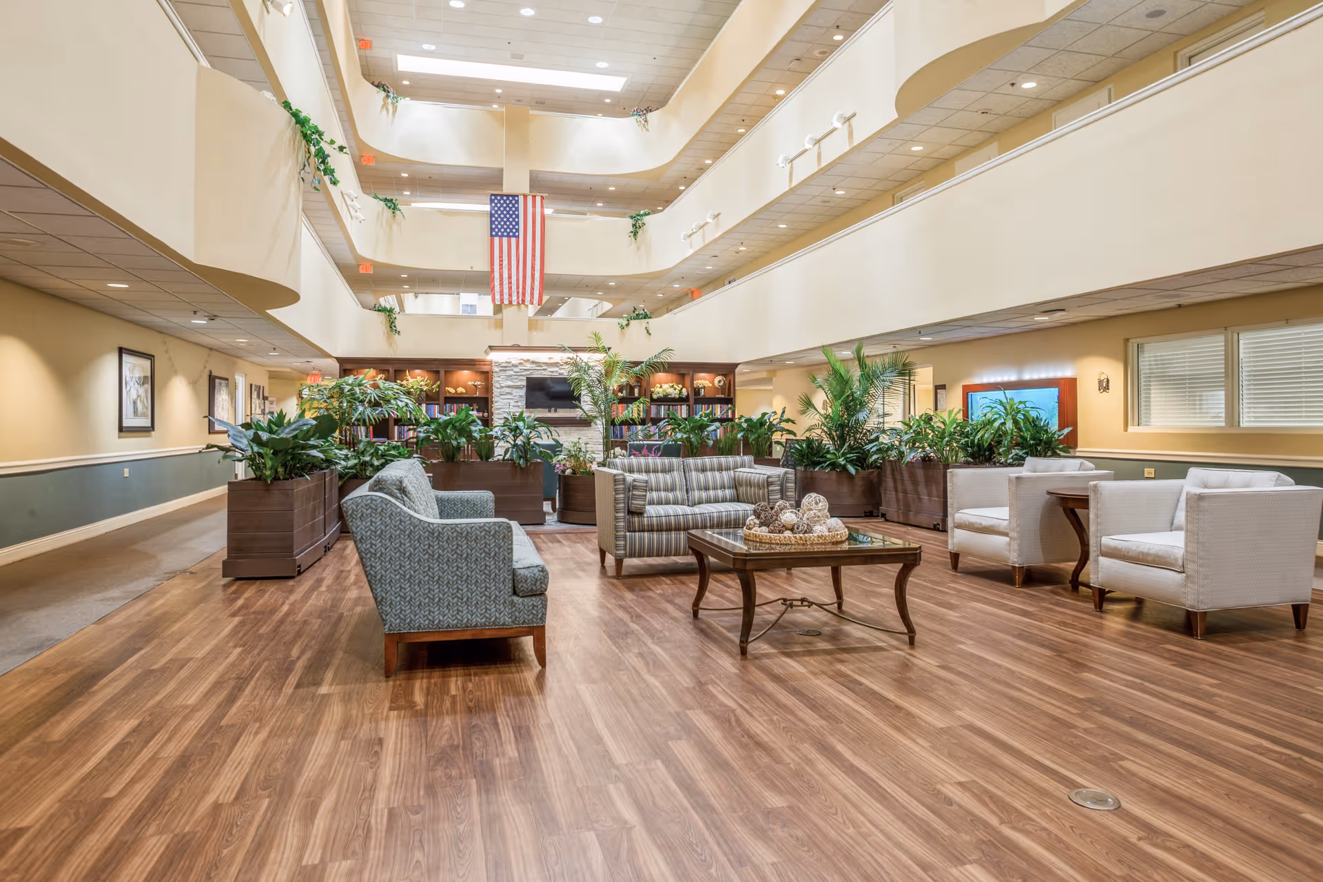Spacious and well-lit common area in a senior living facility with multiple seating arrangements including sofas and armchairs around a wooden coffee table. The room features wooden flooring, large planters with green plants, a stone fireplace with a TV above it, bookshelves, and an American flag hanging from the upper floor balcony. The ceiling is high with multiple levels and recessed lighting.