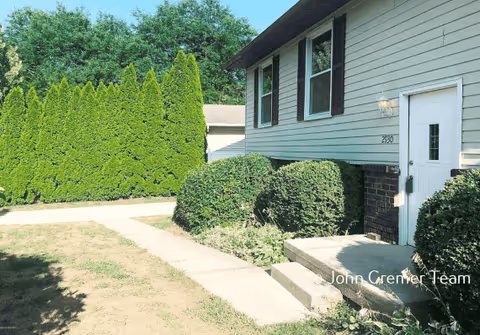 Front exterior of a light-colored house showing a small concrete entry stoop, trimmed bushes, a sidewalk, and tall evergreen trees by the driveway.