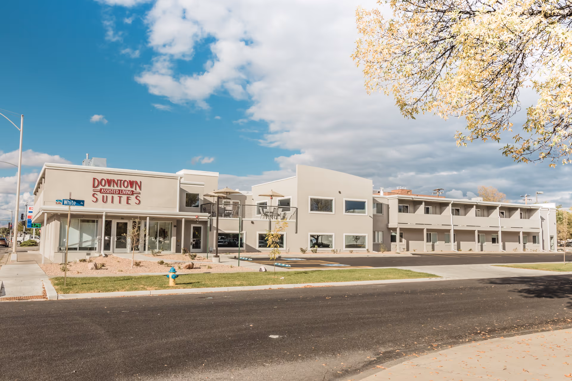 Exterior view of Downtown Assisted Living Suites building on a sunny day with a clear blue sky and some clouds. The building is two stories with balconies and large windows. There is a street and sidewalk in the foreground with a fire hydrant and some small trees and landscaping.