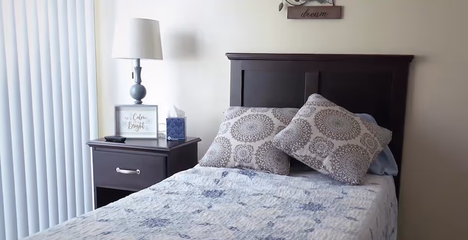 A bedroom with a dark wooden headboard and a bed made with a white and blue patterned quilt and two decorative pillows with circular designs. Next to the bed is a dark wooden nightstand with a lamp, a tissue box, and a framed sign that reads 'All is Calm All is Bright'. Vertical blinds cover the window to the left.