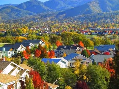 A scenic view of a residential neighborhood with houses surrounded by trees in autumn colors, set against a backdrop of forested hills and mountains under a clear sky.