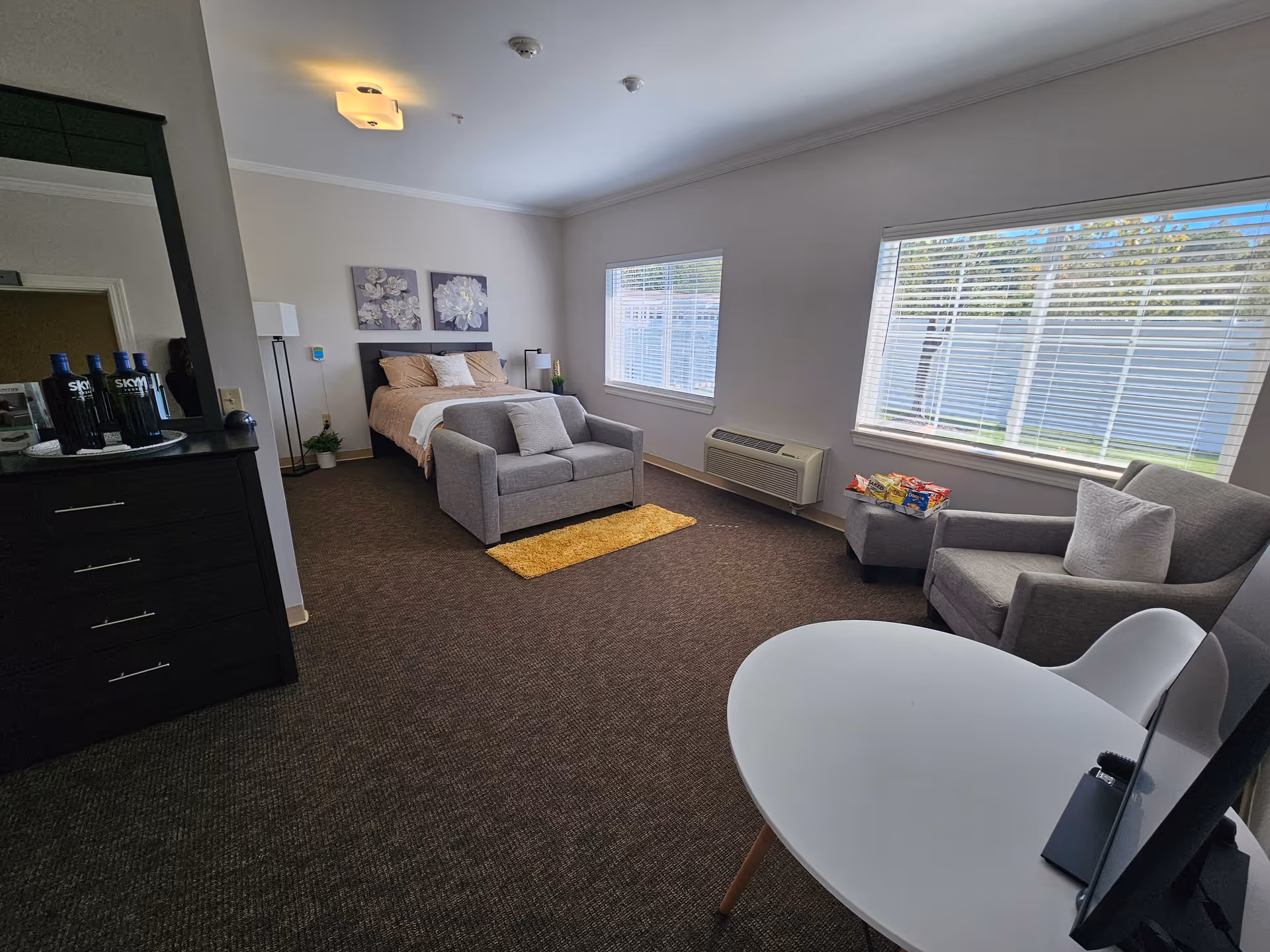 Bright, spacious studio-style bedroom with a bed, loveseat, armchair, dresser and large windows overlooking a yard.