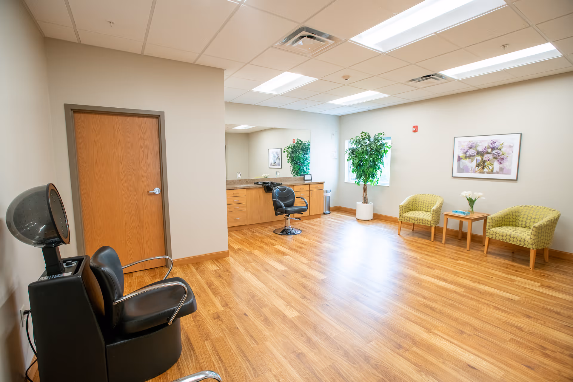 A spacious room with wooden flooring and beige walls featuring a hair salon setup including a black salon chair with a hair dryer, a wooden door, a large mirror above a countertop with drawers, a black swivel chair, two green patterned armchairs with a small wooden table between them holding a vase with white flowers, a large potted plant near a window, and a framed floral picture on the wall.
