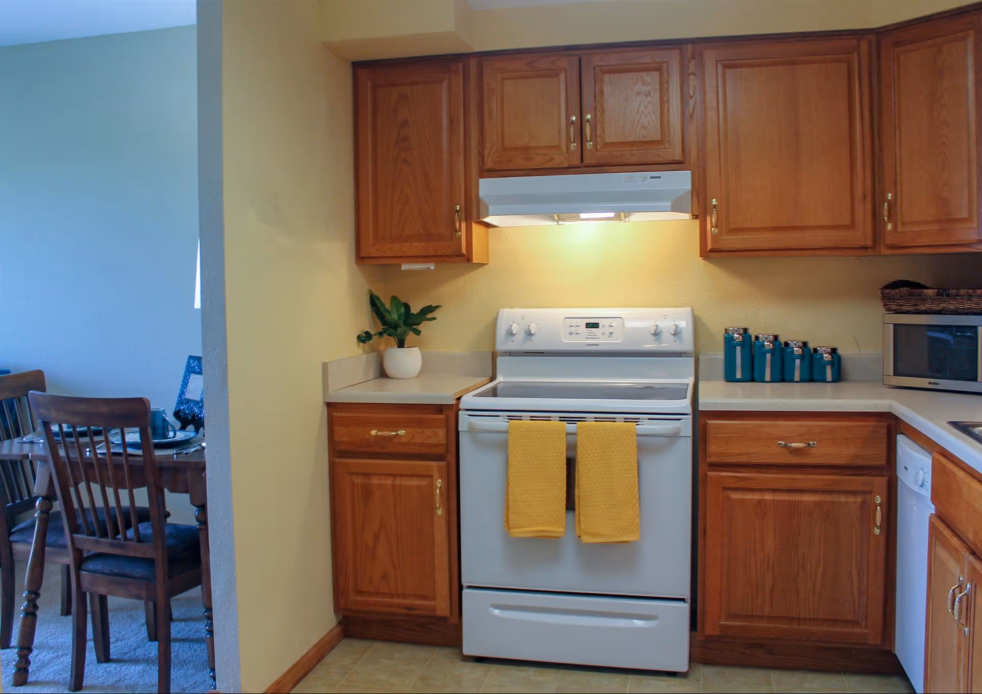 A kitchen with wooden cabinets, a white electric stove with two yellow towels hanging on the handle, a white range hood with a light on, a small potted plant on the left countertop, a set of blue canisters and a microwave on the right countertop. To the left, there is a partial view of a dining area with a wooden table and chairs.