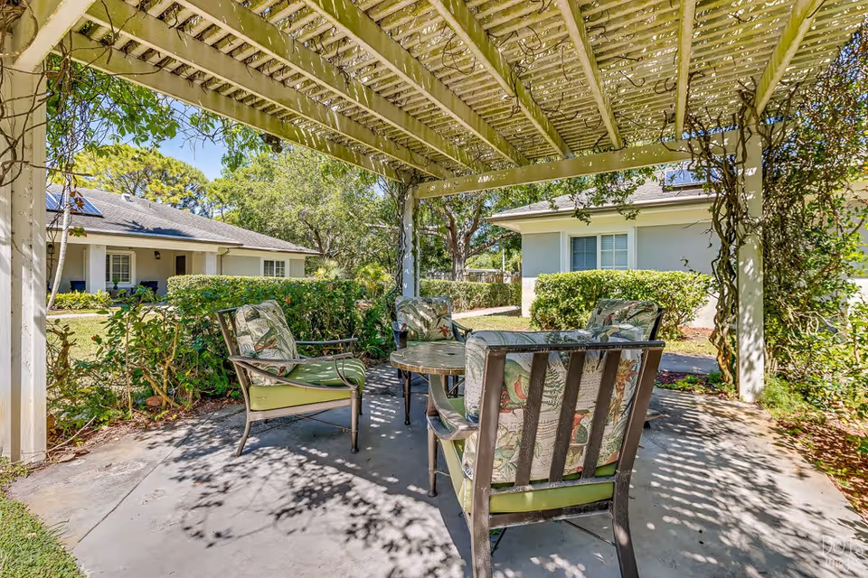 Shaded pergola with a round patio table and four cushioned chairs in a garden courtyard between cottages.