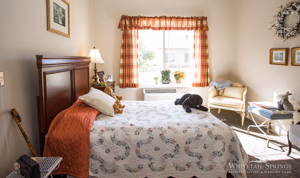 A cozy bedroom with a wooden headboard bed covered with a patterned quilt and pillows, including teddy bears and a black stuffed animal on the bed. There is a window with plaid curtains letting in natural light, a small side table with a lamp and framed photos, a cane leaning against the wall, a cushioned chair, and a small table with books and a decorative rabbit. The walls are decorated with framed pictures and a wreath.