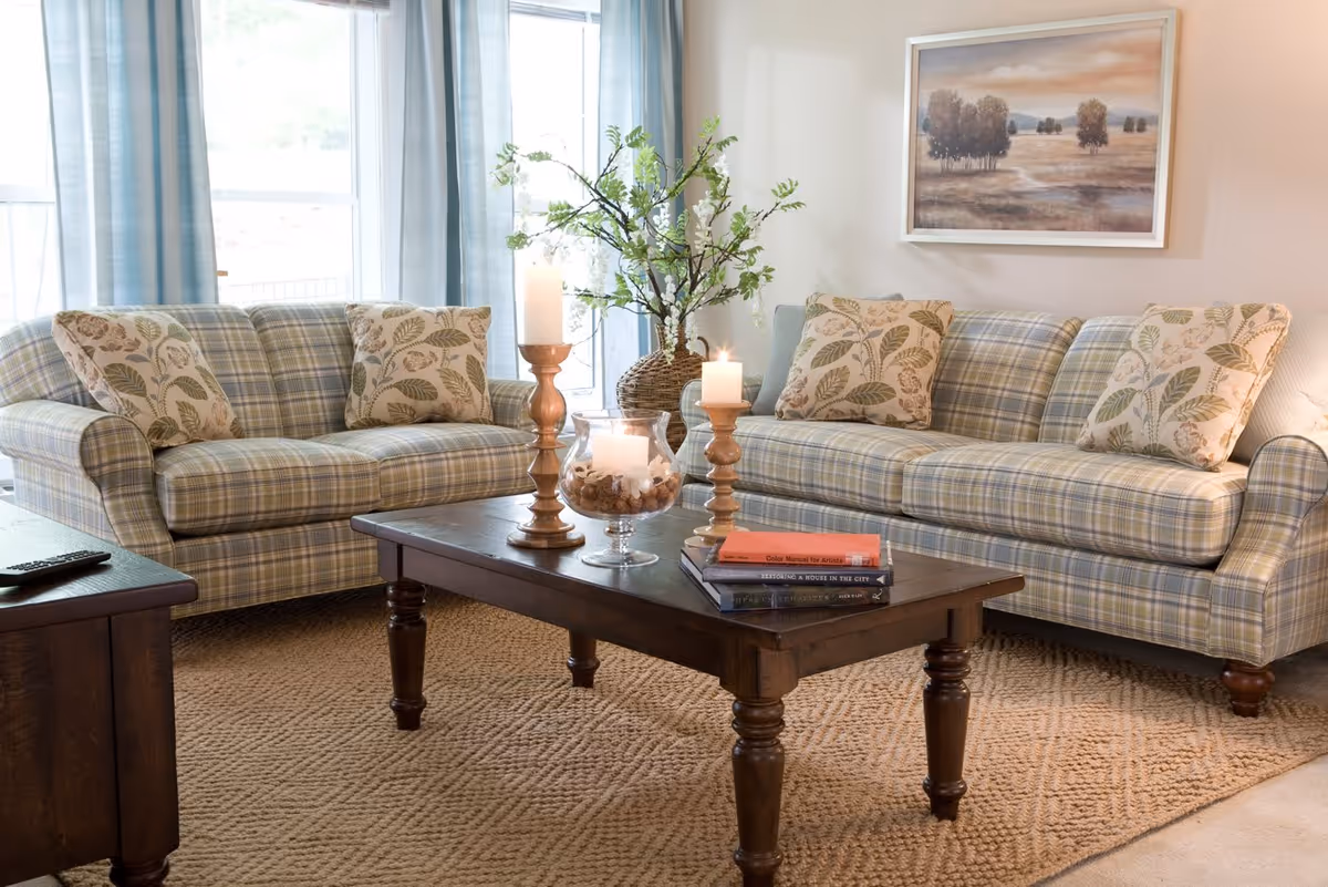 A cozy living room with two plaid sofas adorned with leaf-patterned pillows, a wooden coffee table with candles and books, a large window with light blue curtains, a textured beige rug, and a framed landscape painting on the wall.