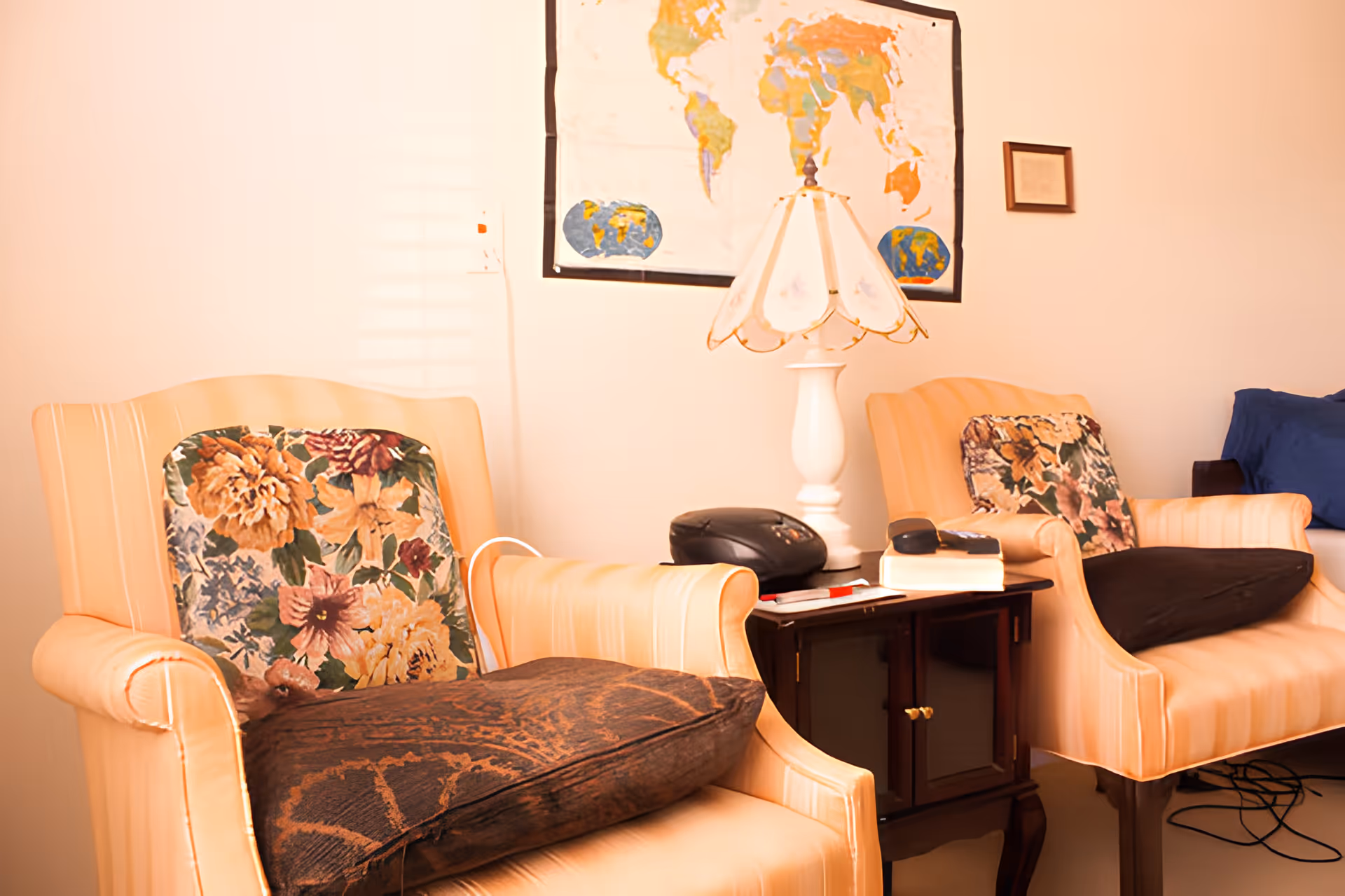 Two beige armchairs with floral and dark cushions are placed on either side of a small dark wooden side table. On the table, there is a white lamp with a decorative shade, a black telephone, a remote control, and a book. A world map and a small framed picture hang on the wall behind the chairs.
