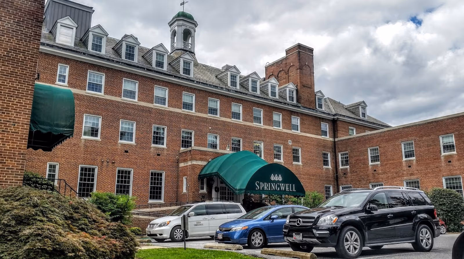 Exterior view of a large brick building with multiple windows and dormers on the roof. The building has a green awning over the entrance with the word 'SPRINGWELL' printed on it. Several cars are parked in front of the building, and there are bushes and greenery near the entrance. The sky is cloudy.