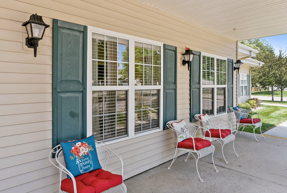 Outdoor covered patio area of a senior living facility with four white metal chairs featuring red cushions and decorative pillows, beige siding with green shutters on windows, and black wall-mounted lantern lights.