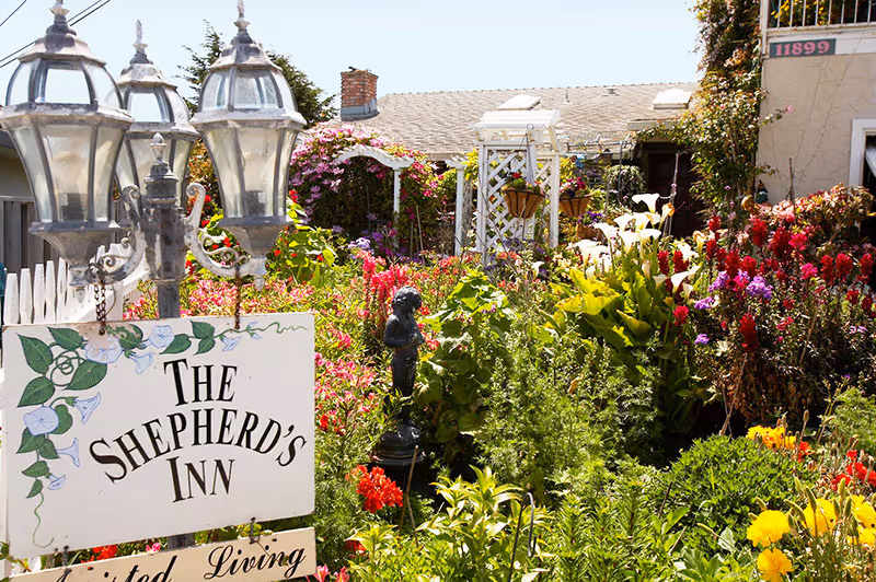 A vibrant garden area at The Shepherd's Inn Assisted Living, featuring a variety of colorful flowers, green plants, a white trellis, and a decorative statue. There are three vintage-style street lamps and a white sign with floral designs that reads 'The Shepherd's Inn'. The building and roof are visible in the background under a clear sky.