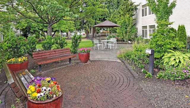 A landscaped courtyard with brick pavers, wooden benches, colorful flower planters, and a patio table with an umbrella beside a senior living building.