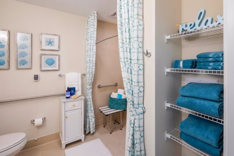 A clean and well-organized bathroom featuring a walk-in shower with a patterned curtain, a small white cabinet with toiletries on top, a toilet with a toilet paper holder, and wall art depicting sea-themed images. On the right side, there are shelves stocked with neatly folded teal towels and a decorative 'relax' sign on the top shelf.