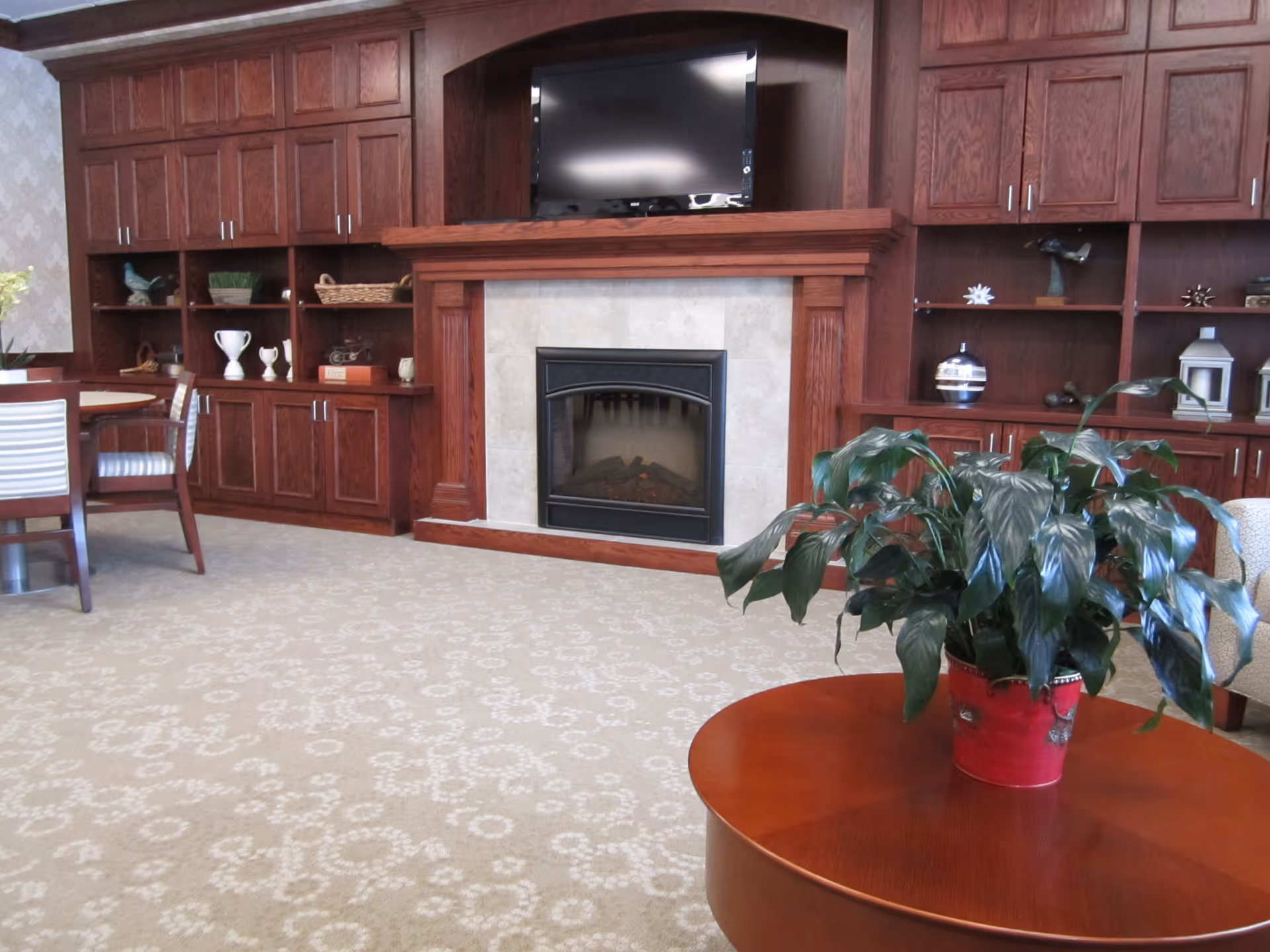 A cozy common room with built-in wooden cabinets and a fireplace, a TV above, and a potted plant on a round table in the foreground.