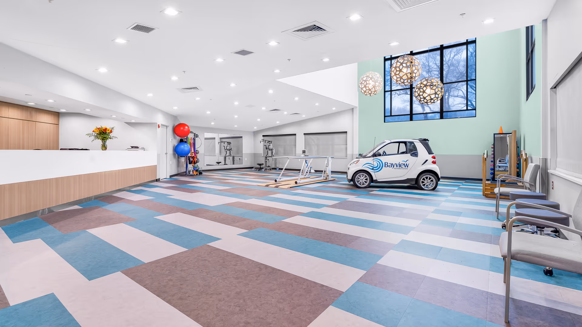 Spacious bright rehabilitation therapy room with a patterned floor, reception desk on the left, exercise equipment and a small Bayview-branded car near large windows.