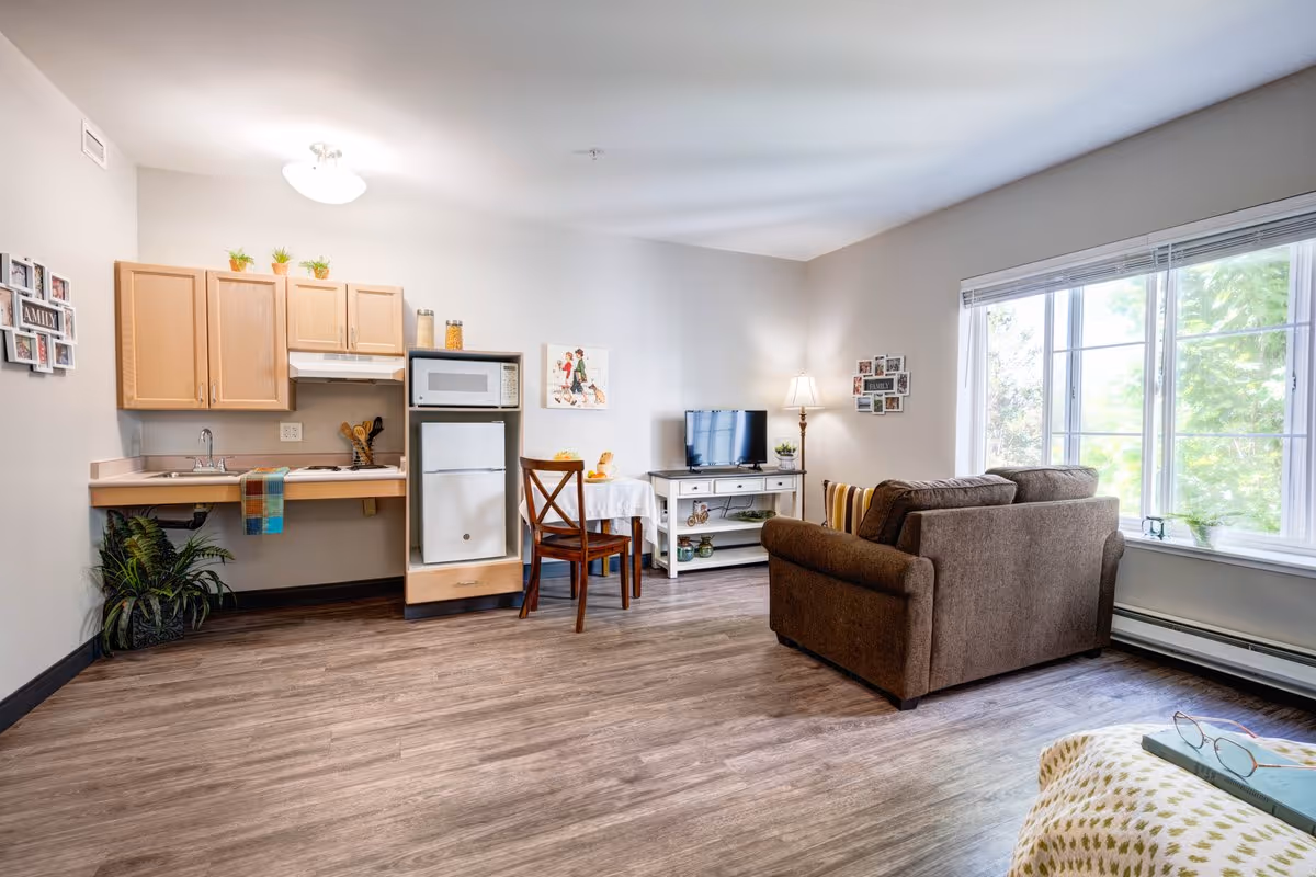 A bright and cozy living space featuring a small kitchen area with wooden cabinets, a microwave, and a refrigerator. There is a small dining table with two chairs next to the kitchen. A brown sofa faces a TV on a white stand, with a floor lamp beside it. Large windows allow natural light to fill the room, and the floor is covered with wood-style flooring. Decorative plants and framed photos adorn the walls and surfaces.