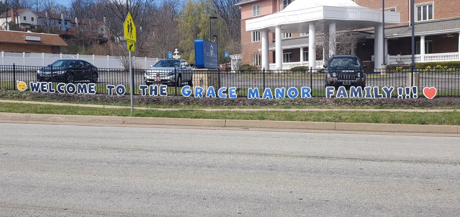 Outdoor view of Grace Manor at North Park showing the front of the building with a black fence in front. Large letters attached to the fence spell out 'WELCOME TO THE GRACE MANOR FAMILY!!!' with an emoji of a smiling face and a red heart at the ends. Several cars are parked behind the fence and trees and houses are visible in the background.