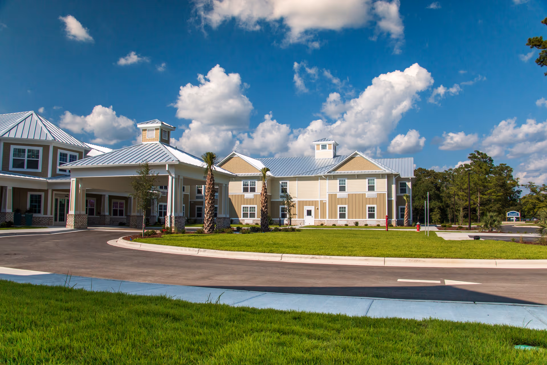 Exterior view of Arbor Landing at Hampstead senior living facility showing a large building with beige and white siding, metal roofing, and a covered entrance. The scene includes a curved driveway, green lawn, palm trees, and a bright blue sky with scattered clouds.