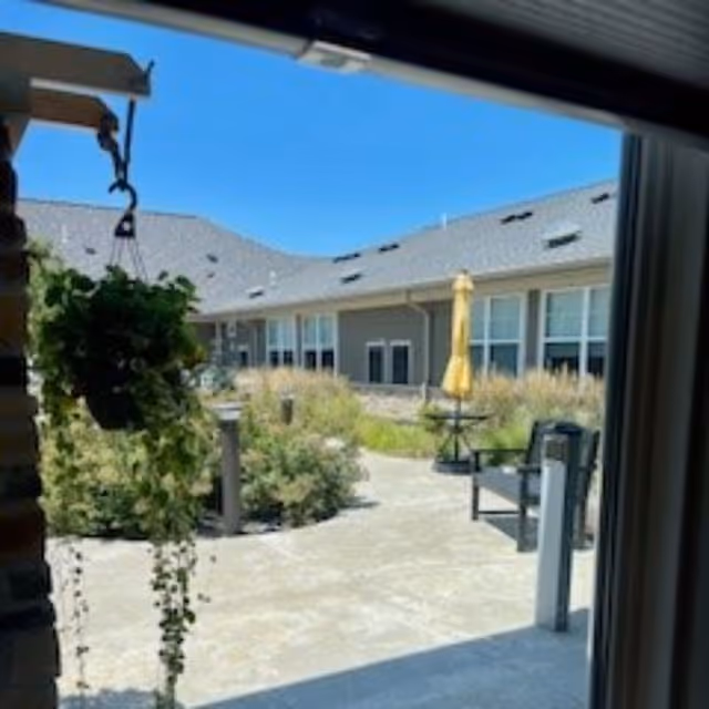 View through a doorway into a sunny courtyard with planted beds, patio seating, and a yellow umbrella outside a single-story senior living building.