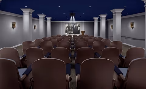A conference or presentation room with multiple rows of brown cushioned chairs facing a podium and a large black and white photograph on the front wall. The room has white columns, wall sconces, and a dark blue ceiling with recessed lighting.
