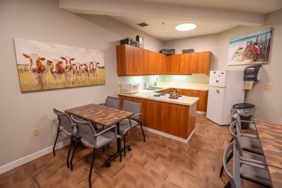 A small kitchen and dining area with wooden cabinets, a white refrigerator, microwave, and a kettle on the stove. There are two dining tables with gray chairs. The walls are decorated with a large picture of cows on the left and a beach scene with a red bicycle on the right.