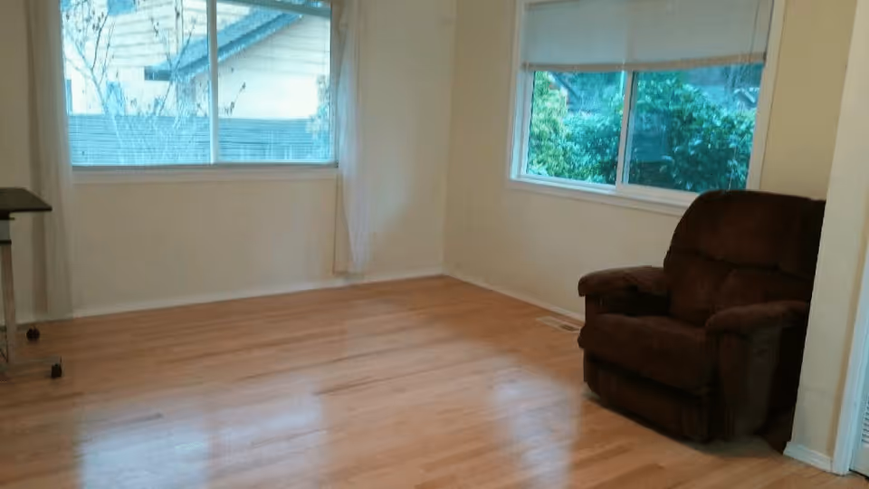 A small room with light-colored walls and wooden flooring featuring two large windows with views of greenery outside. There is a single dark brown cushioned armchair positioned near one window and a small table with wheels partially visible on the left side.