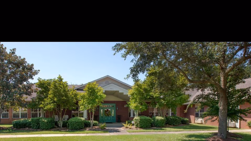 Front exterior view of Knollwood Pointe Assisted Living and Memory Care building with a green entrance door, surrounded by trees and bushes under a clear blue sky.