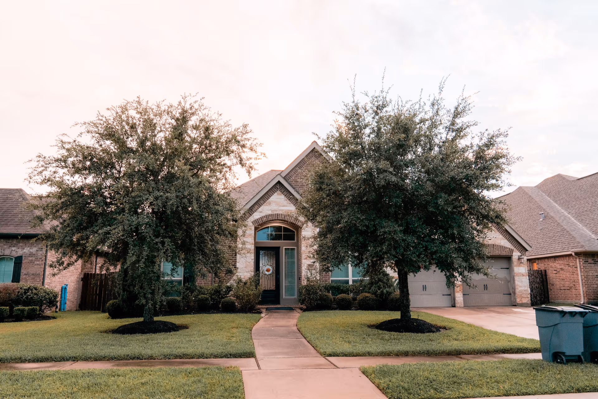Front exterior view of a single-story brick house with a stone arch around the front door, two large trees in the front yard, a concrete pathway leading to the entrance, and a double garage on the right side.