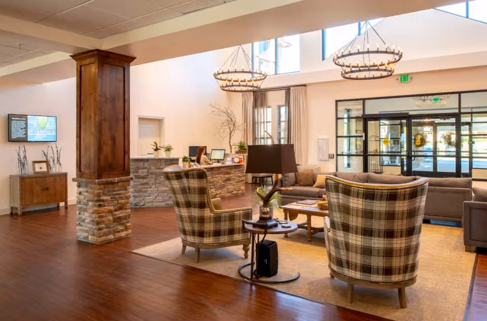 A spacious and well-lit senior living facility lobby with wooden flooring, stone and wood pillar, a reception desk with a staff member, comfortable seating area including plaid armchairs and a gray sofa, large windows, and modern chandeliers hanging from the ceiling.