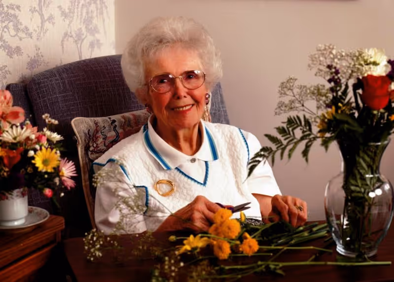 An elderly woman with white hair and glasses is sitting at a table arranging yellow flowers. She is smiling and wearing a white sweater vest over a collared shirt. There are various flowers in vases on the table and nearby, and a patterned pillow is visible on the chair behind her.