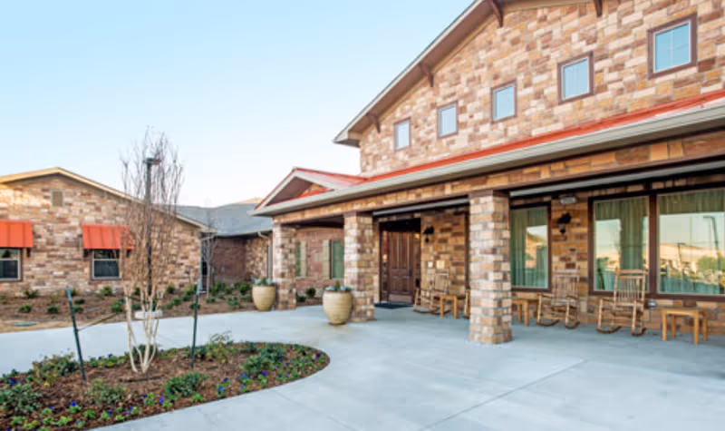 Exterior view of a senior living facility with a brick facade, a covered entrance with rocking chairs, large windows with curtains, and a landscaped area with plants and small trees.
