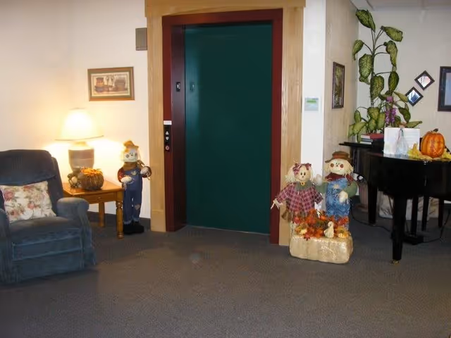 Interior view of a senior living facility common area featuring a green elevator door framed in wood. To the left, there is a blue armchair with a floral pillow and a wooden side table with a lamp and a pumpkin decoration. To the right of the elevator, there are two scarecrow decorations on a hay bale, a black piano with plants and framed pictures on the wall behind it.