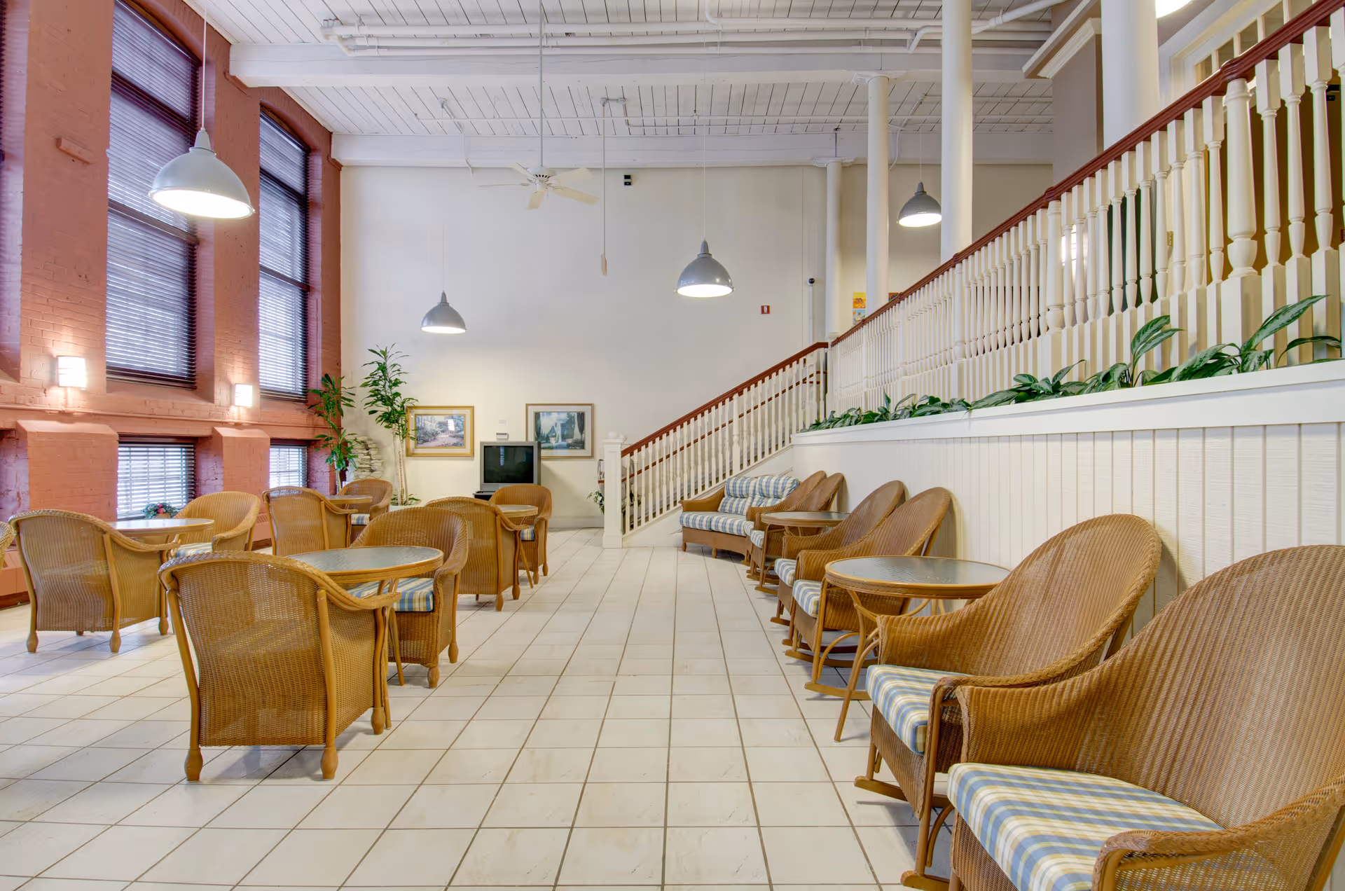 A spacious indoor common area with wicker chairs and small round tables arranged in rows. The room has large windows with blinds on the left side, white tiled flooring, and a staircase with white railings on the right. There are hanging pendant lights and framed pictures on the far wall.