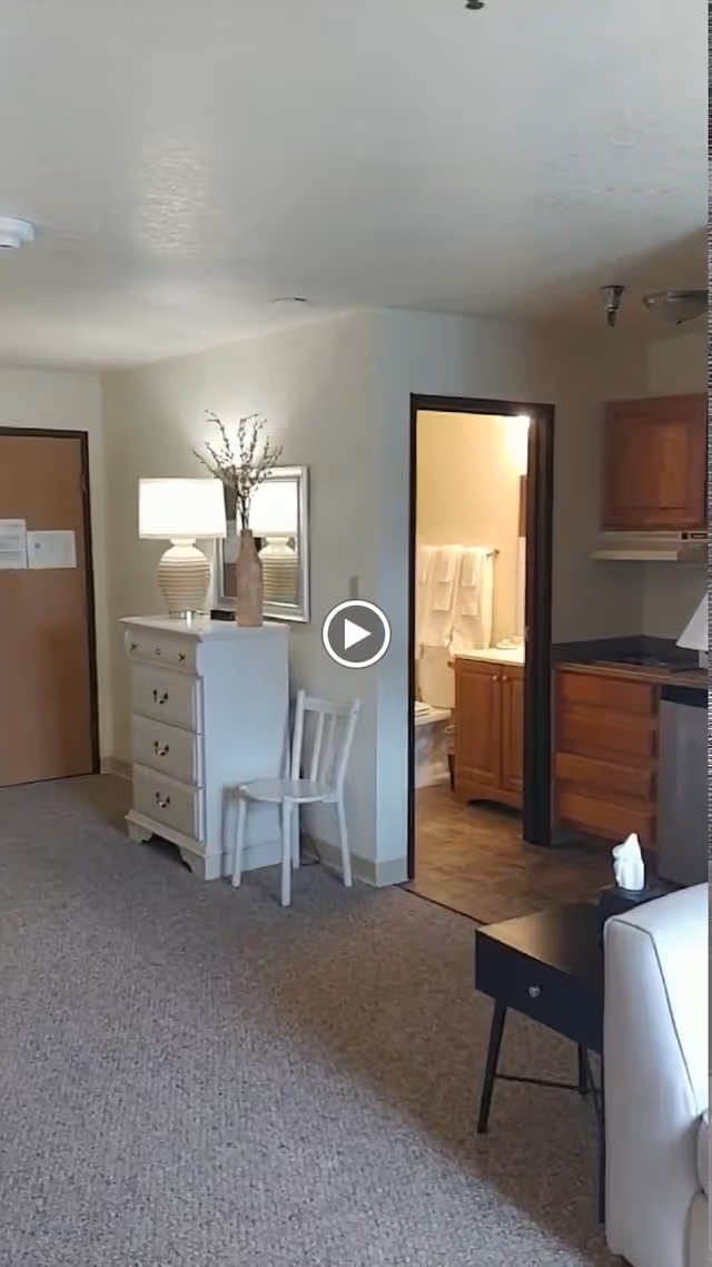 Interior view of a senior living facility room showing a white dresser with a lamp and a vase on top, a white chair next to it, an open doorway leading to a bathroom with towels hanging, and part of a kitchenette with wooden cabinets and a black countertop. A small black side table with a tissue box is visible next to a white armchair.