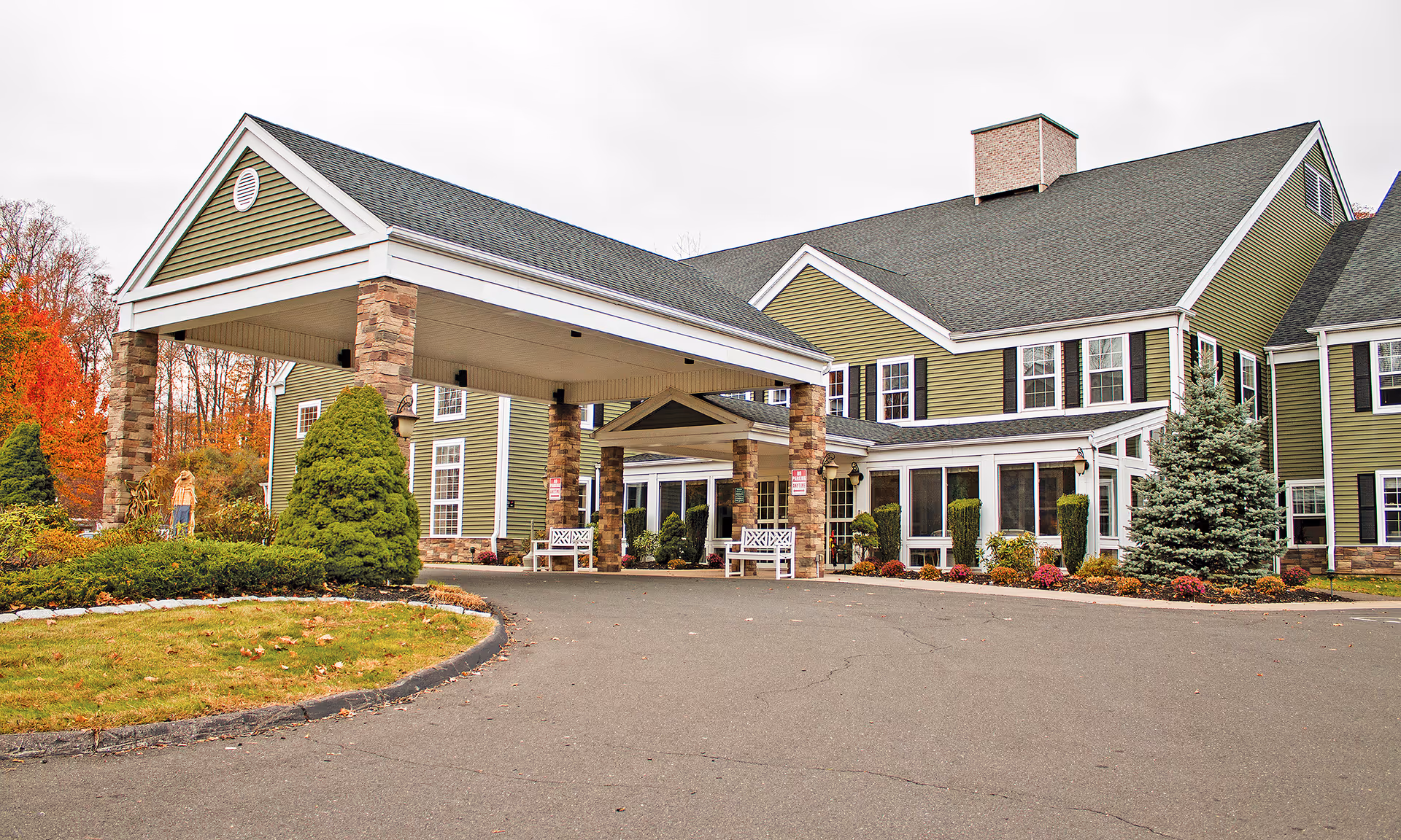 Exterior view of a senior living facility named The Residence at Ferry Park, featuring a covered entrance with stone pillars, green siding, multiple windows, landscaped bushes, and trees with autumn foliage.