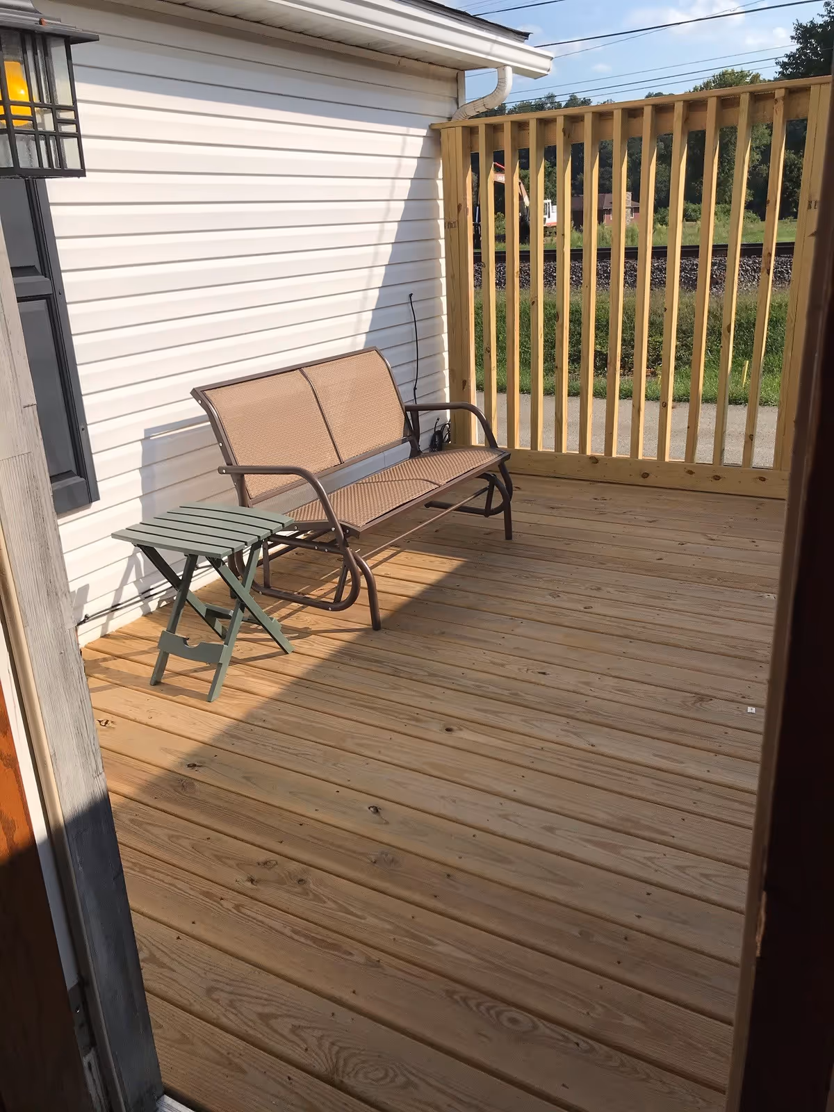 A wooden deck attached to a white-sided building with a brown metal bench and a small green folding table. The deck has a wooden privacy fence with vertical slats and a view of greenery and a railroad track in the background.