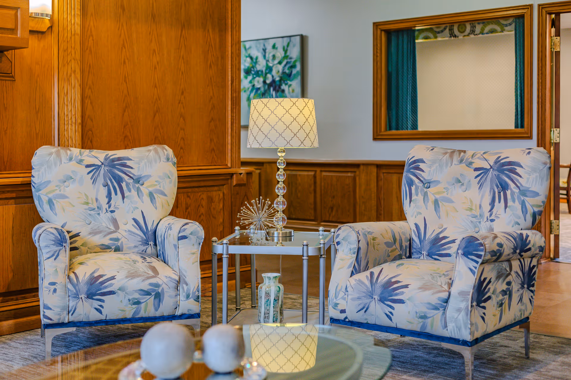 Two floral-upholstered armchairs flanking a glass-top side table with a lamp in a wood-paneled sitting area.