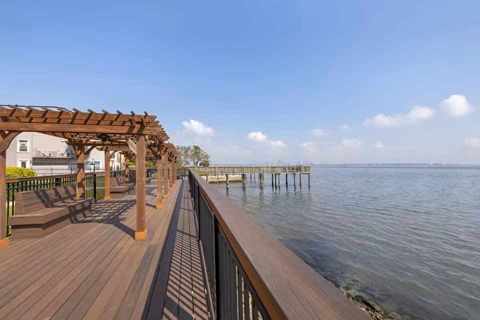 Outdoor wooden deck area with pergolas and benches overlooking a large body of water under a clear blue sky.