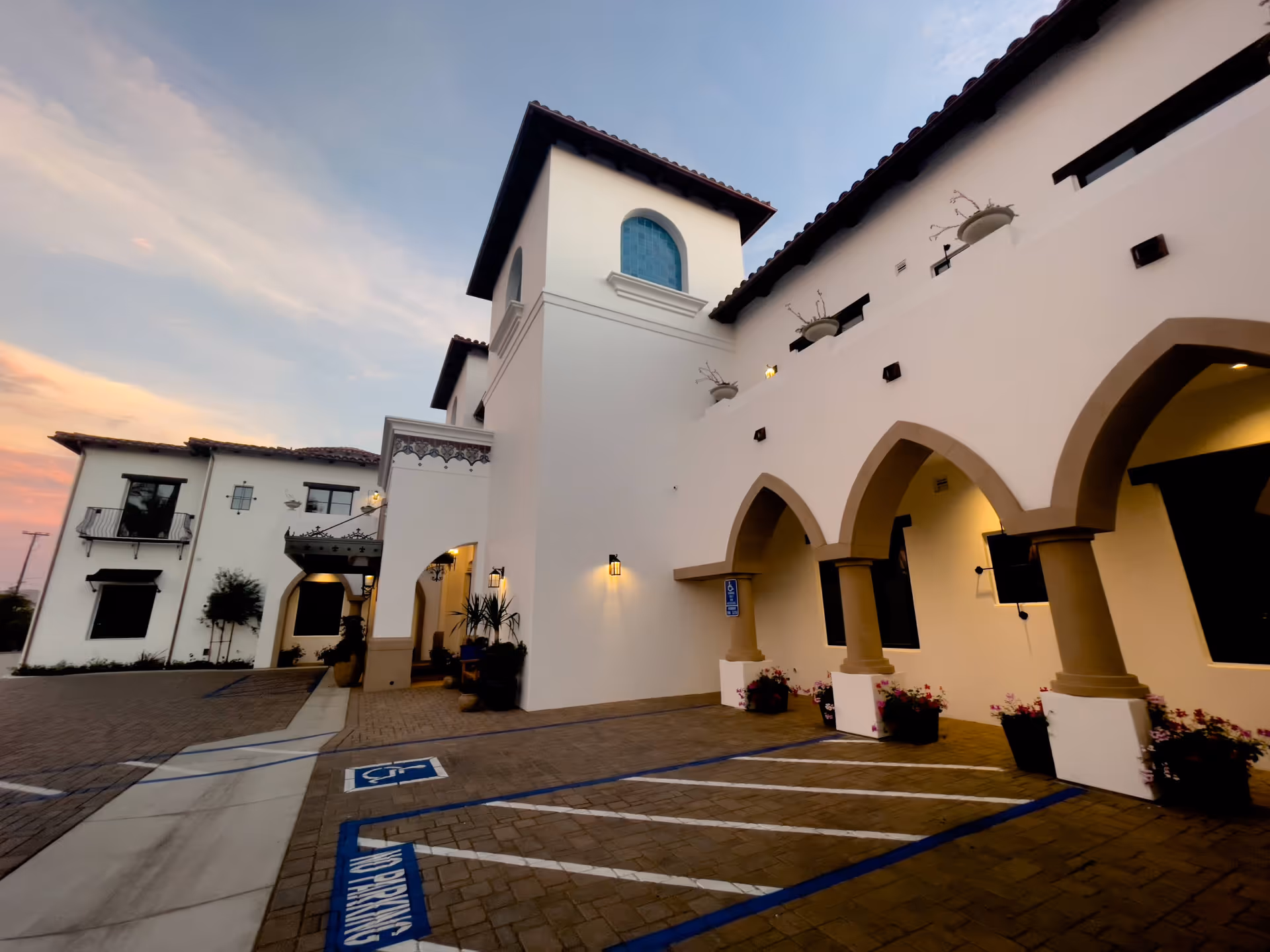 Exterior view of a white building with Spanish-style architecture at sunset, featuring arched doorways, a tower with a blue-tiled window, and a parking area with handicapped parking spaces and no parking zones.