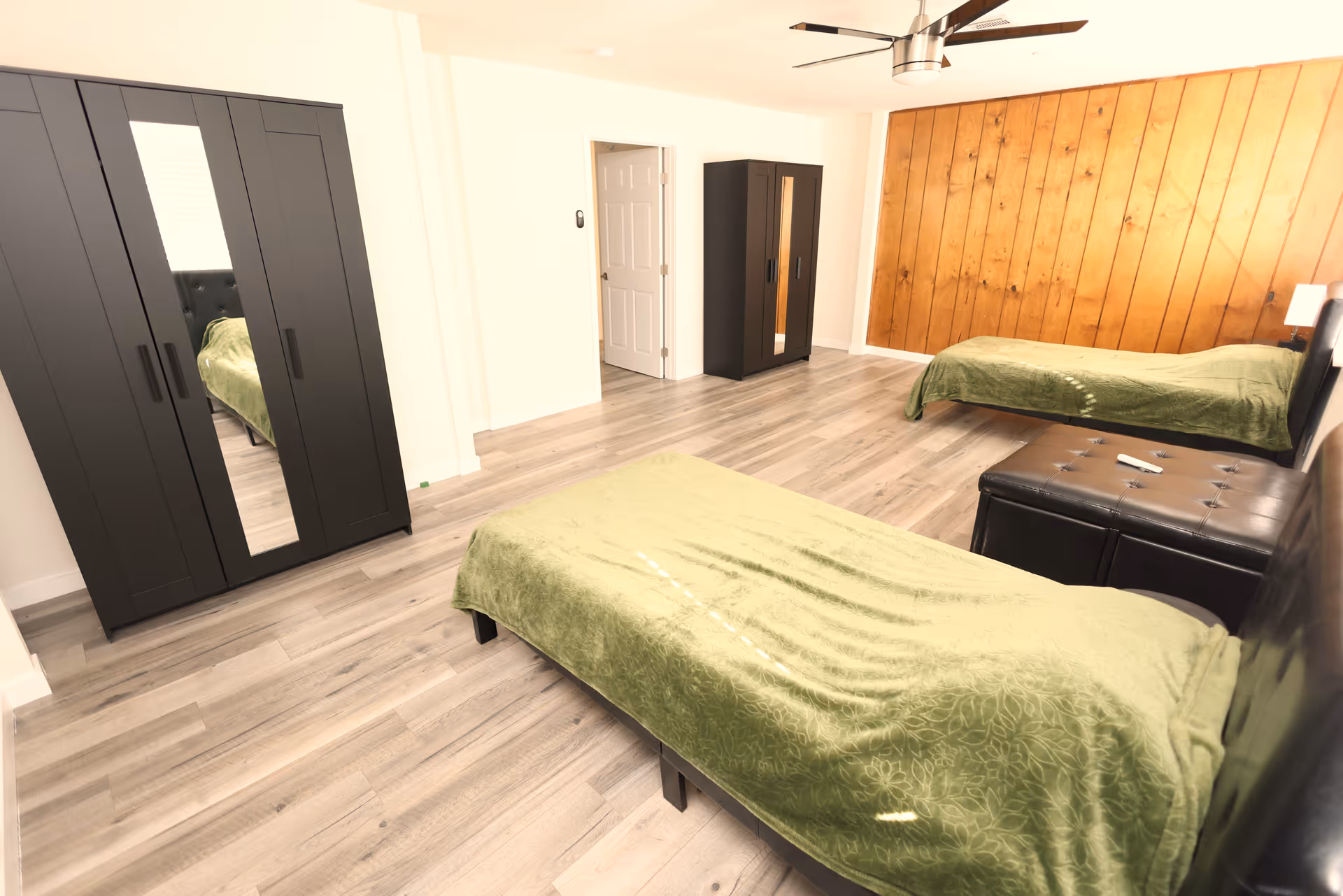 Spacious bedroom with two twin beds covered in green blankets, dark wardrobes, wood-paneled accent wall, and light wood flooring.