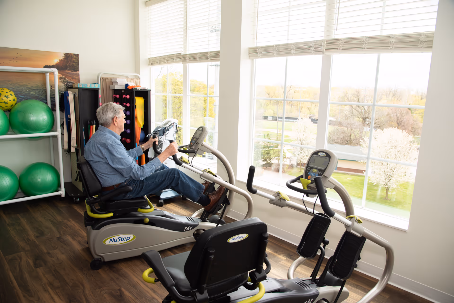An elderly man exercising on a NuStep recumbent cross trainer in a bright room with large windows overlooking trees and a grassy area. The room has wooden flooring and exercise equipment including green exercise balls and resistance bands on a rack.