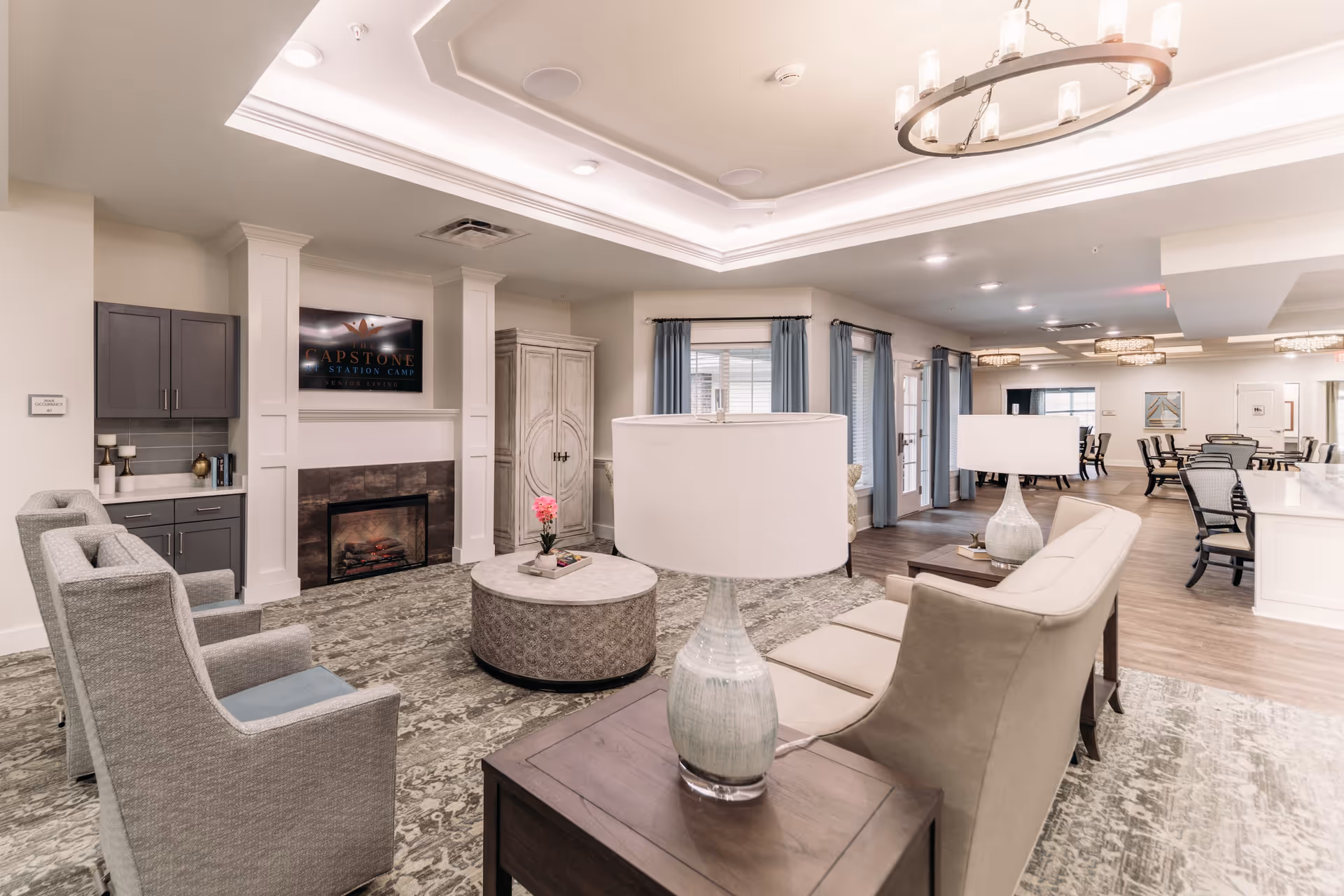A spacious and well-lit living room area in The Capstone at Station Camp featuring comfortable seating including armchairs and sofas arranged around a round coffee table. There is a fireplace with a TV mounted above it displaying the facility's name. The room has light-colored walls, a patterned carpet, and large windows with blue curtains. In the background, there is a dining area with tables and chairs, and modern ceiling light fixtures.