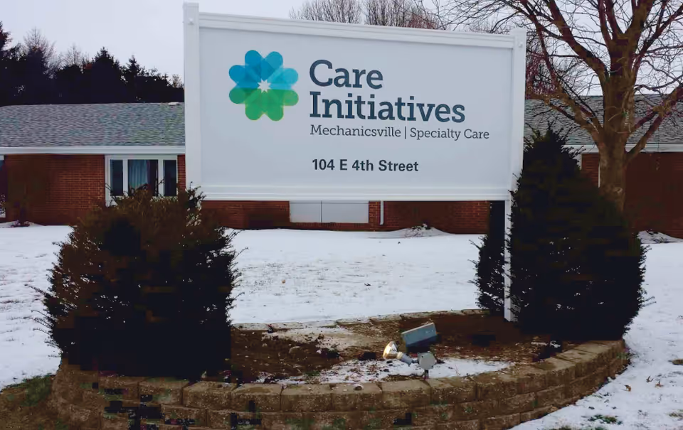 Outdoor view of a sign for Care Initiatives Mechanicsville Specialty Care located at 104 E 4th Street, surrounded by snow-covered ground, bushes, and a brick building in the background.