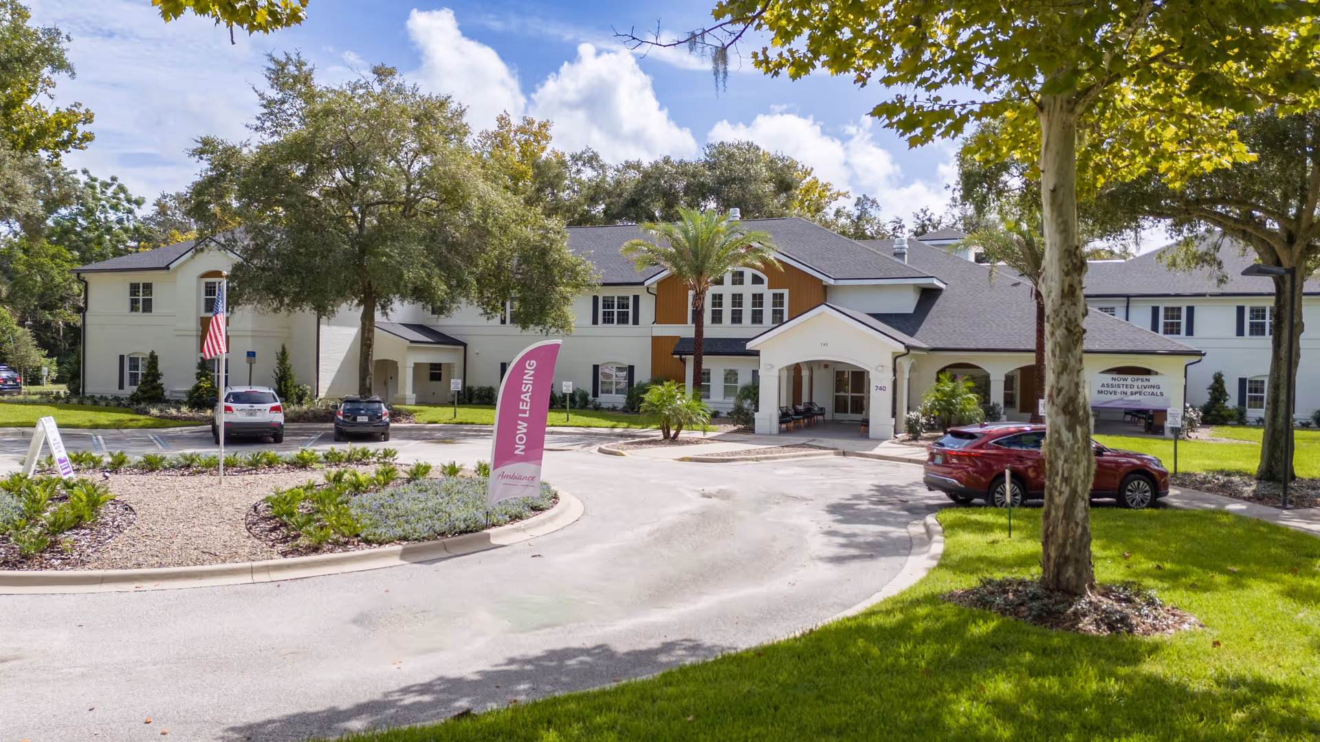 Front exterior view of Ambiance at Maitland, a two-story assisted living facility with a circular driveway, landscaped greenery, palm trees, and several parked cars. There are signs indicating 'Now Leasing' and 'Now Open Assisted Living Move-In Specials'.