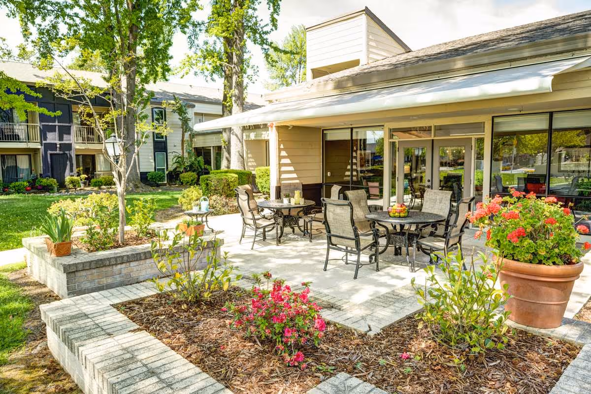 Outdoor patio area at Greenhaven Place with several metal tables and chairs arranged on a paved surface. The patio is surrounded by landscaped garden beds with various plants and flowers, including a large potted plant with red flowers. In the background, there are trees and a two-story building with balconies and large windows.