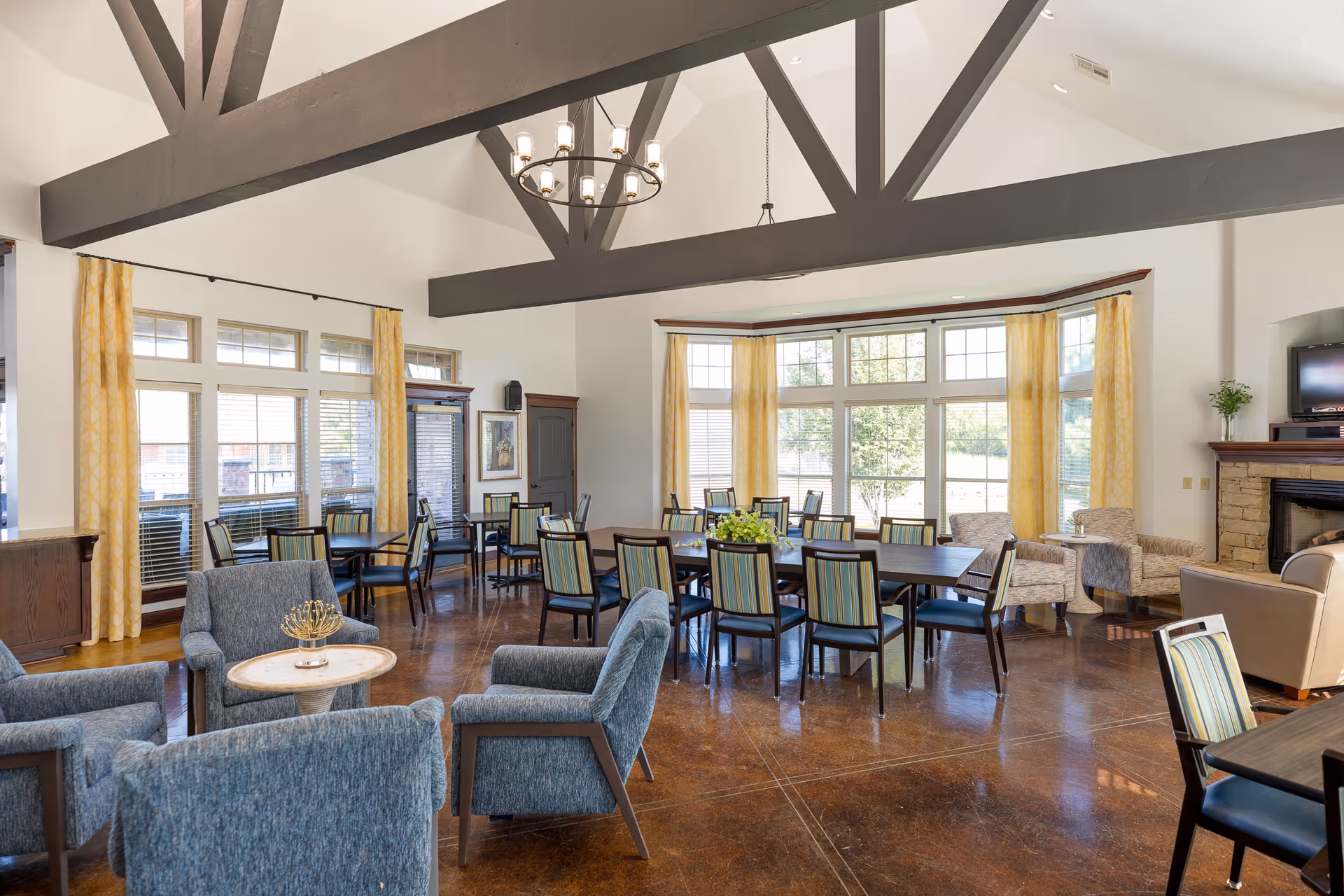 A spacious and well-lit common area in a senior living facility with high vaulted ceilings featuring exposed dark wooden beams. The room has large windows with yellow patterned curtains allowing natural light to fill the space. There are multiple seating arrangements including blue upholstered armchairs around a small round table, and several dining tables with striped cushioned chairs. A stone fireplace with a TV on top is visible on the right side, and the floor is a polished brown surface.