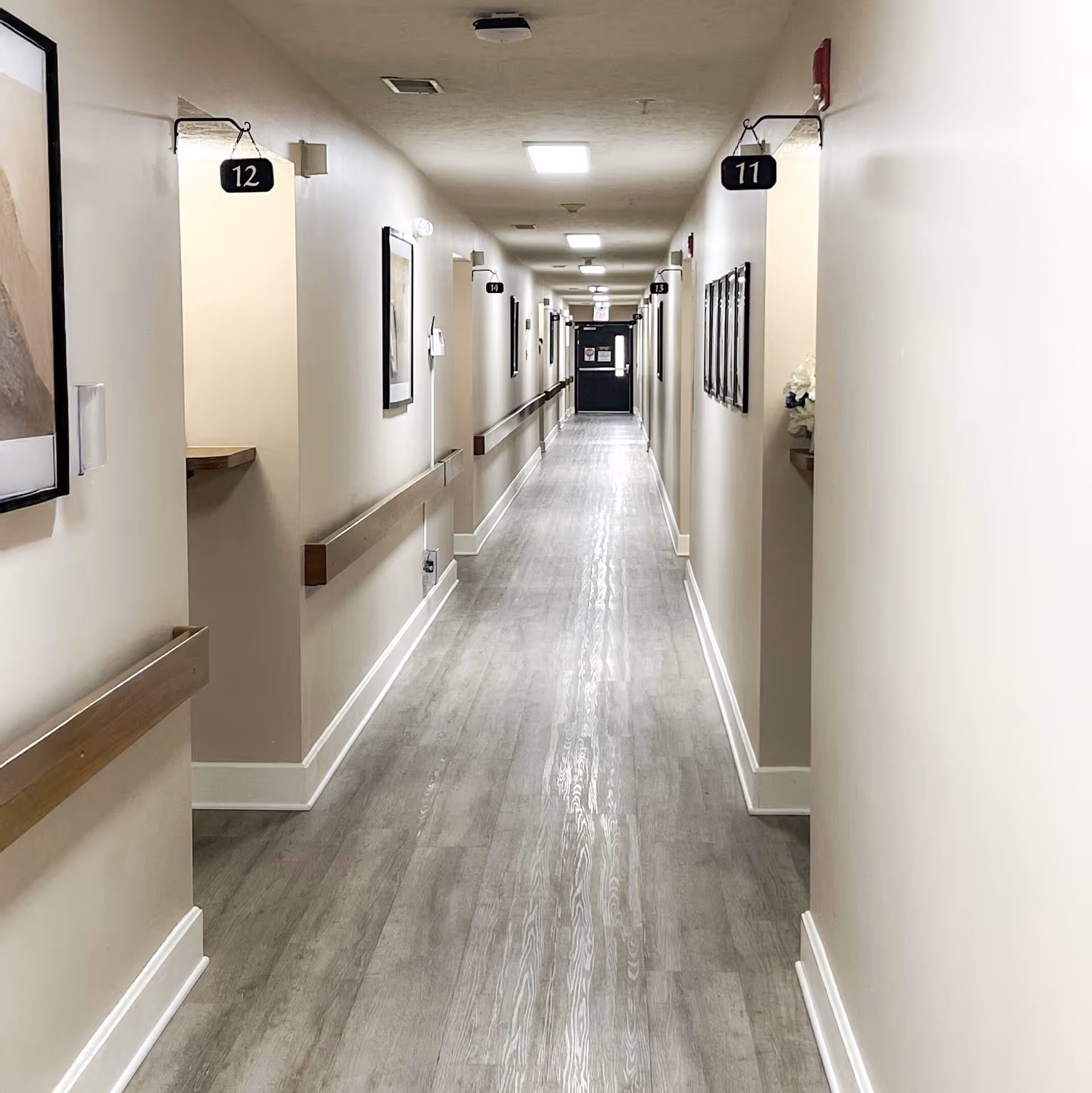 A long, well-lit hallway in a senior living facility with light-colored walls and wood-patterned flooring. The hallway has handrails on both sides and numbered room signs hanging above each doorway. Several framed pictures are mounted on the walls, and a door is visible at the far end of the corridor.
